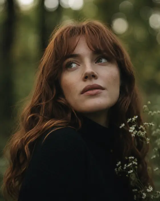 Create a cinematic portrait of a woman in her late 20s with wavy auburn hair and soft bangs, her face turned up and to the side with a relaxed, thoughtful look. She is wearing a black turtleneck sweater. The background is a softly blurred forest with rich green tones and round bokeh from light filtering through the trees, with small white wildflowers near her shoulder. The camera is at eye level, slightly below her face looking up. Use soft, diffused natural light with muted earthy tones and a gentle film grain. Shot with an 85mm lens, very shallow depth of field. Use aspect ratio 4:5.