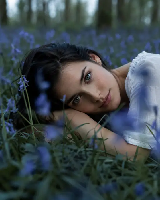 Create a cinematic portrait of a woman with dark hair resting her head on her arm while lying in a field of bluebell flowers in a forest. She is wearing a simple white top and looking directly into the camera with a soft, open expression. Blades of grass and out-of-focus blue flowers fill the foreground and frame her face. The camera is at ground level, low among the flowers with a shallow focus on her eyes. Use cool, slightly desaturated green and blue tones with soft overcast light for a moody, romantic feel. Shot with a 50mm lens. Use aspect ratio 4:5.