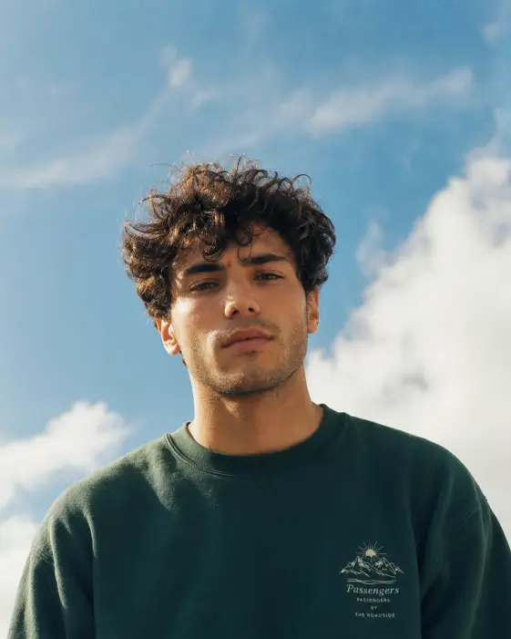 Create a cinematic portrait of a young man with tousled dark curly hair and light stubble standing outdoors, looking straight into the camera with a calm, neutral expression. He is wearing a dark green crewneck sweatshirt with a small printed graphic on the chest. The background is a bright blue sky with soft white clouds. The camera is at a slightly low angle looking up at him. Use clean natural daylight with warm skin tones against the cool blue sky and a subtle vintage film grade. Shot with a 35mm lens. Use aspect ratio 4:5.