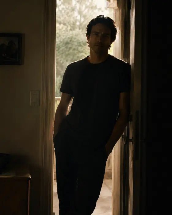 Create a cinematic portrait of a man standing in an open doorway, leaning one shoulder against the frame with his hands in his pockets and a calm, unreadable expression. He is wearing a plain dark t-shirt and trousers. Bright light spills in from behind him, rimming his shoulders and hair while leaving his face in soft shadow. The interior around him is dim and muted. The camera is at eye level, framing him from the knees up. Use strong backlight with warm highlights and deep shadow for a quiet, filmic mood. Shot with a 35mm lens. Use aspect ratio 4:5.