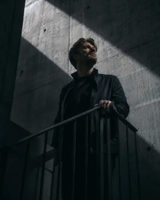 Create a cinematic portrait of a man standing on a concrete stairwell landing, one hand on the metal railing as he looks up toward a shaft of light. He is wearing a fitted dark jacket. Hard light cuts through the space, throwing sharp diagonal shadows across the walls and his face. The background is raw concrete in cool grey tones. The camera is at a low angle looking up the stairwell. Use high-contrast directional light with cool shadows and a gritty cinematic grade. Shot with a 35mm lens. Use aspect ratio 4:5.