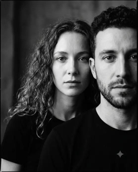 Create a black and white close-up portrait of a couple with their faces layered, the man in the front right of the frame partially cropped and the woman standing just behind his shoulder on the left, both looking directly into the camera with calm, intense expressions. The man has short dark curly hair and a trimmed beard, and the woman has long loose curly hair falling around her face. Both wear plain dark shirts. Shoot at eye level, extremely tight framing so their faces fill the frame edge to edge. Soft directional studio light with fine film grain and deep contrast. Moody editorial photography style. Use aspect ratio 4:5.